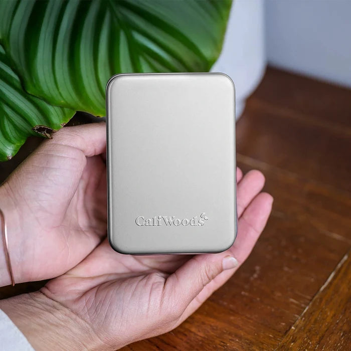 Hand holding a small metallic device with 'CaliWoods' branding, against a wooden surface and plant background.