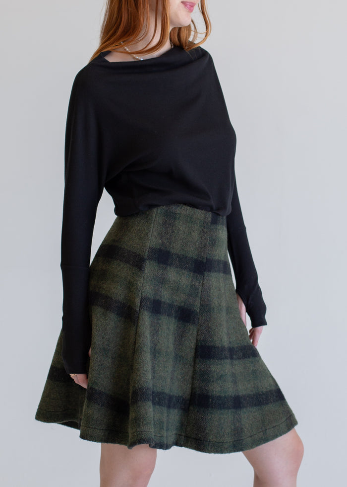 Person wearing a black long-sleeve top and green plaid skirt against a plain background