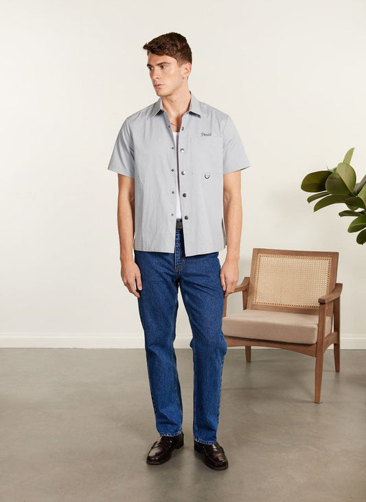 Full body shot of a model wearing the Percival Auxiliary Boxy Clerk Shirt in light blue with dark jeans and brown loafers.