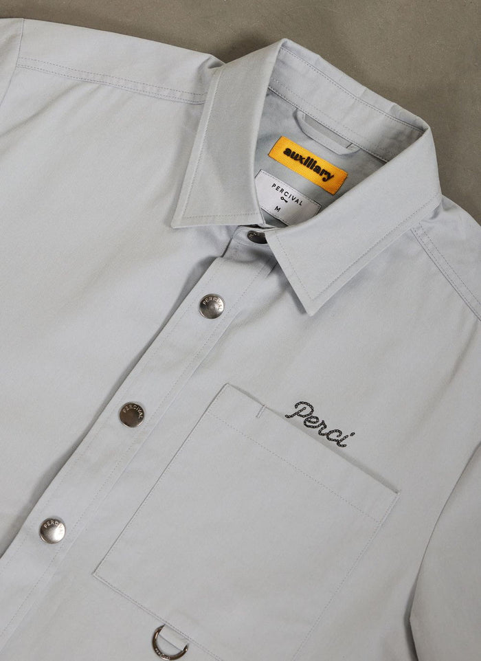 A close-up of the light blue Percival Auxiliary Boxy Clerk Shirt, showing the 'Perci' embroidery and branded snap buttons.