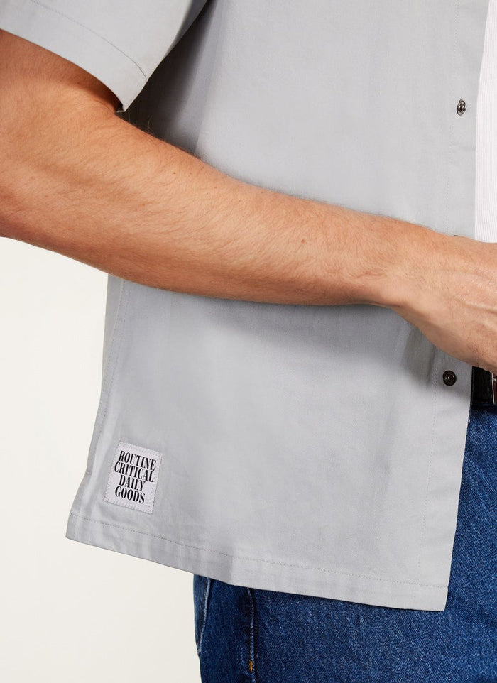A close-up of a model wearing the Percival Auxiliary Boxy Clerk Shirt in light blue, showing the embroidered hem patch.