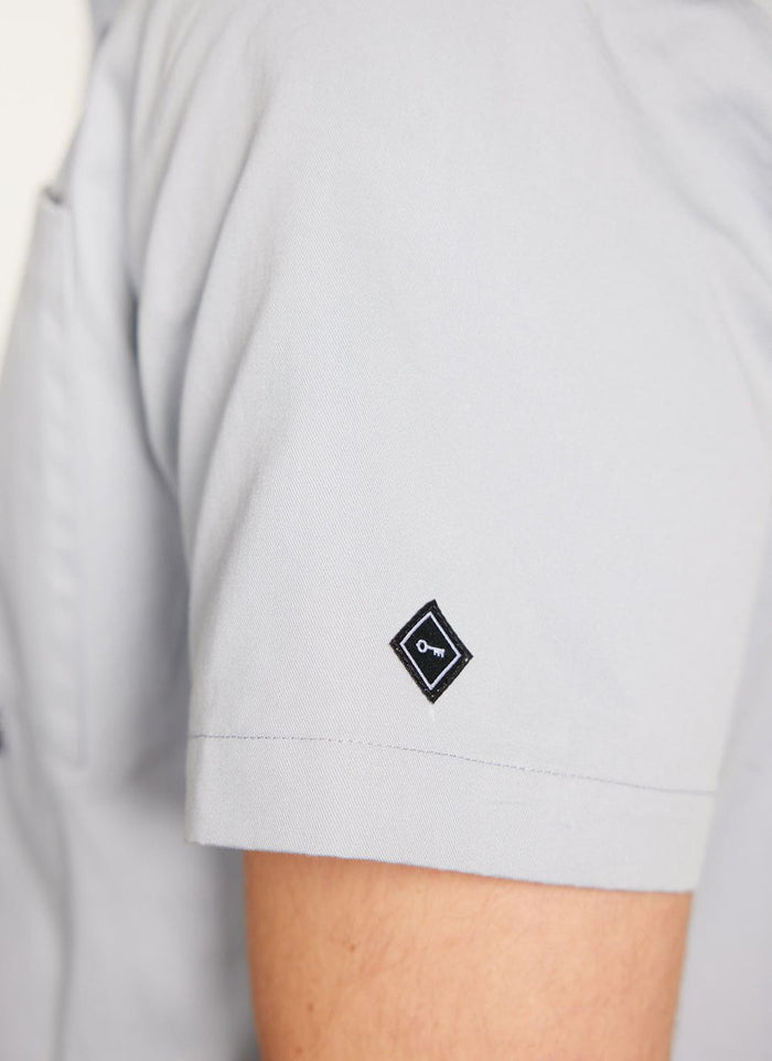 Close up of the light blue Percival Auxiliary Boxy Clerk Shirt showing the embroidered key logo on the short sleeve.