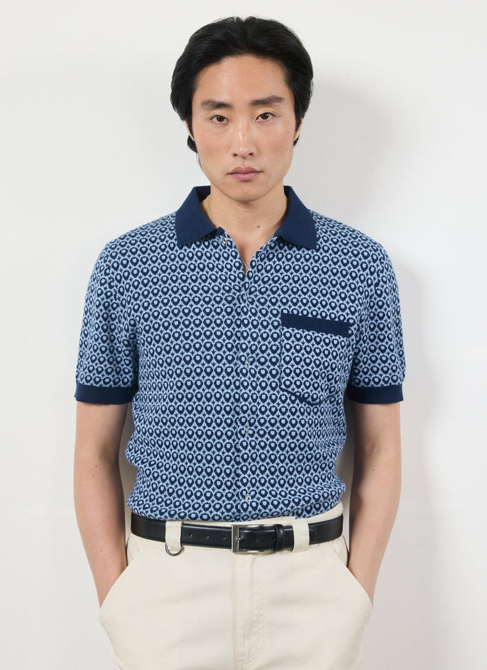 Model wears the Percival Casa Piccante Shirt, a blue jacquard knitted polo, with cream trousers against a white wall.