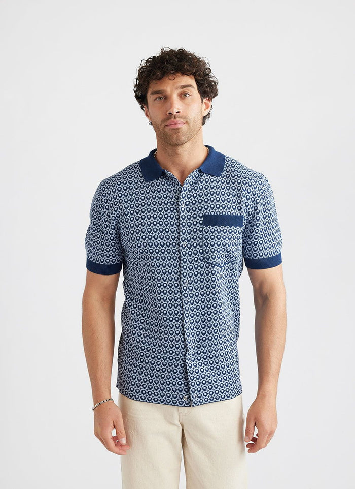 A model wears the Percival Casa Piccante knitted shirt in blue jacquard with cream trousers against a white background.