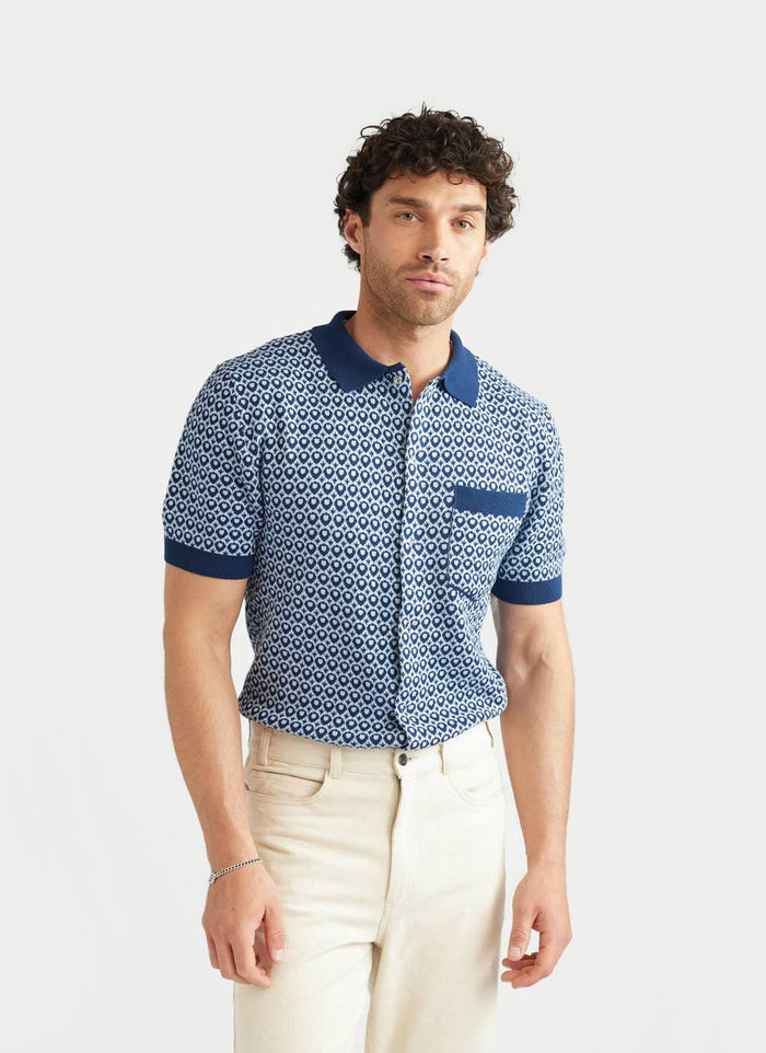 A model wears the Percival Casa Piccante short-sleeved knitted shirt in a blue jacquard pattern with cream trousers.