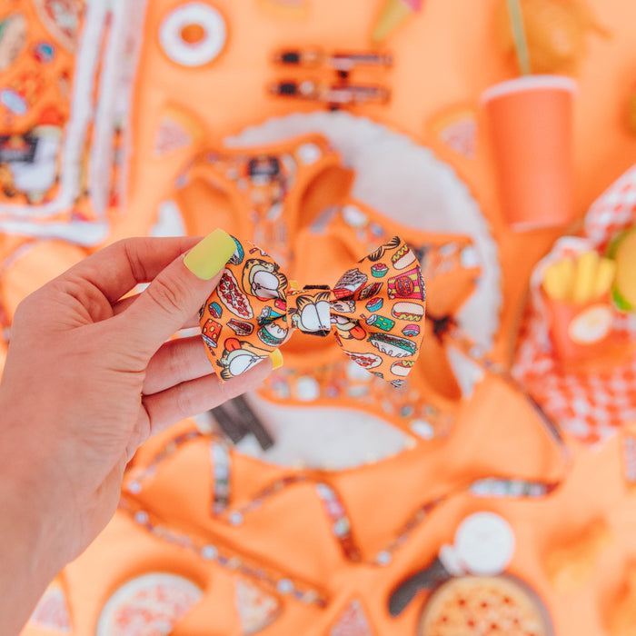 A hand with neon yellow nail polish holds the Sassy Woof Cat Bowtie - Garfield™, featuring playful food illustrations; the background displays orange-themed snacks and accessories.