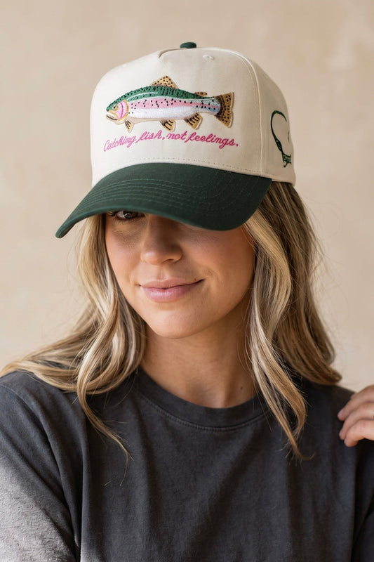 Cream and forest green trucker hat featuring an embroidered rainbow trout with the phrase "Catching Fish, Not Feelings" and an adjustable mesh back.
