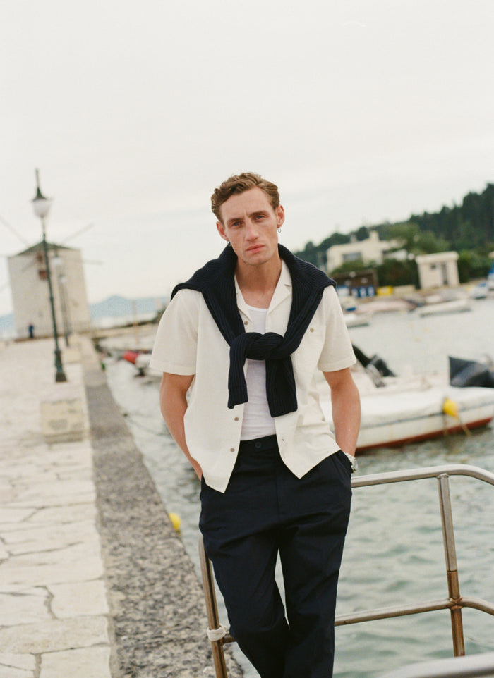 Percival cream linen short sleeve cuban shirt styled with a navy jumper over the shoulders at a coastal harbour.