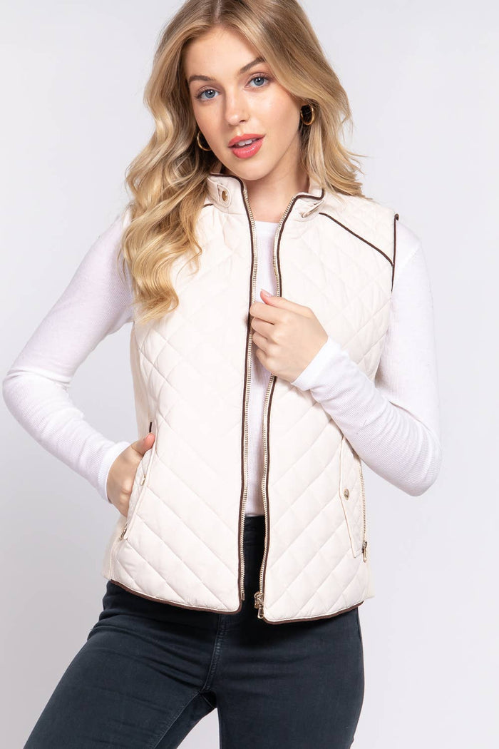 A female model wearing a beige diamond quilted vest with suede piping details and ribbed sleeves, paired with a white long-sleeve top.
