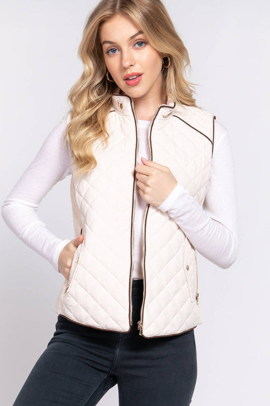 A female model wearing a beige diamond quilted vest with suede piping details and ribbed sleeves, paired with a white long-sleeve top.