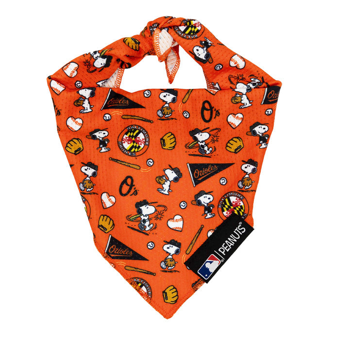 The Sassy Woof Dog Bandana - MLB™ x Peanuts™ Baltimore Orioles™ features Snoopy playing baseball, team logos, gloves, bats, and hearts on orange fabric. It has a knotted top with PEANUTS and MLB tags on the side.