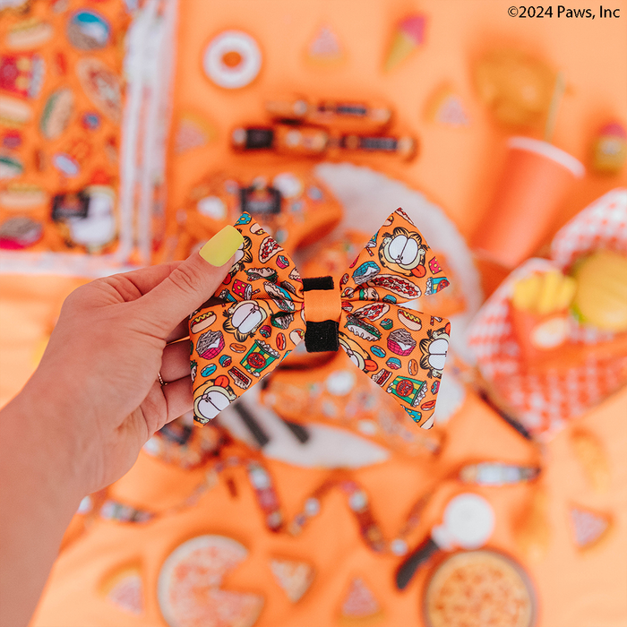 A hand with yellow-green nail polish holds the Sassy Woof Dog Sailor Bow - Garfield™, featuring playful food illustrations, matching an adjustable harness set; assorted orange snacks decorate the orange background.
