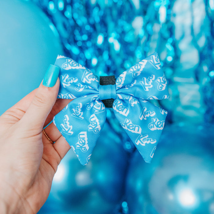 A hand with blue-painted nails holds the Sassy Woof Dog Sailor Bow - KEN™, patterned with “Ken” in white. Blue balloons, a waste bag holder, and metallic blue streamers create a festive background.
