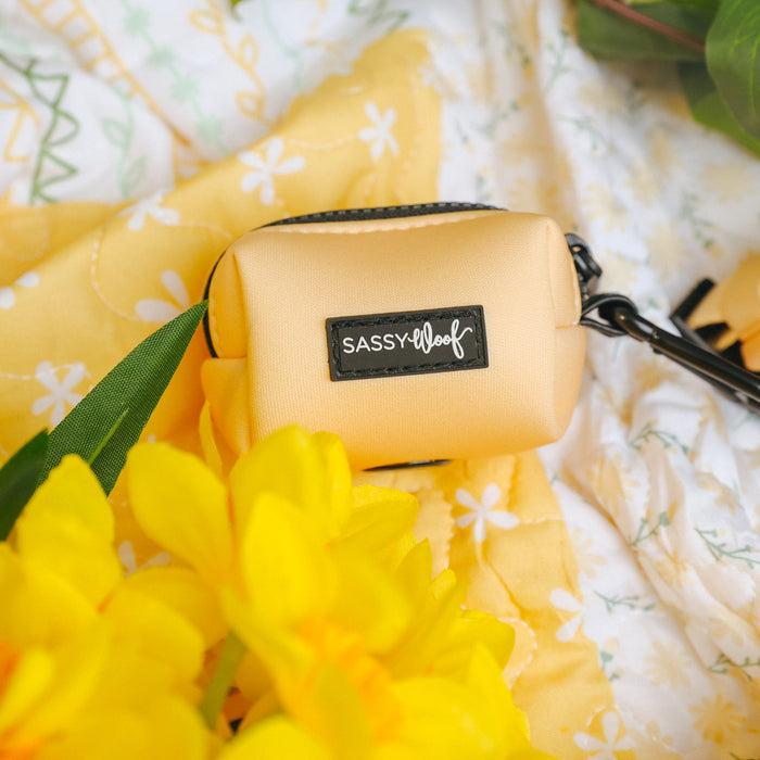 The Sassy Woof Dog Waste Bag Holder - Daffodil, in yellow neoprene, sits on a yellow and white floral fabric with bright yellow flowers and green leaves—a chic accessory for dog walks.