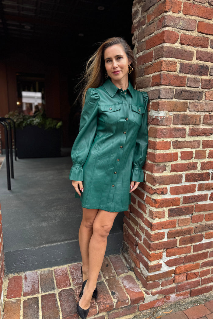 Woman in a green leather dress standing against a brick wall.