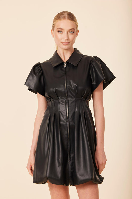 Model in the Dolce Cabo Vegan Leather Zip Bubble Dress - Black. A black faux leather mini dress with a structured fit, short puff sleeves, a pointed collar, and a full zip front. The skirt has a soft flare for a polished, modern silhouette.