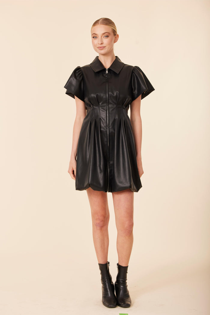 Model in the Dolce Cabo Vegan Leather Zip Bubble Dress - Black. A black faux leather mini dress with a structured fit, short puff sleeves, a pointed collar, and a full zip front. The skirt has a soft flare for a polished, modern silhouette.
