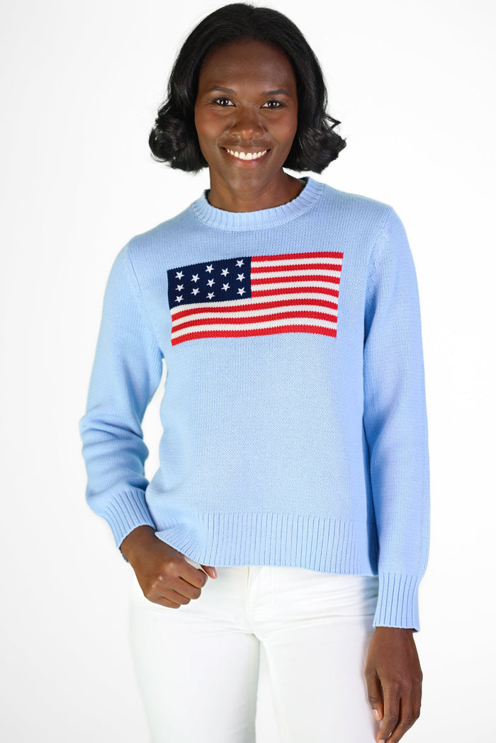Smiling model in a light blue sweater with American flag, paired with white pants