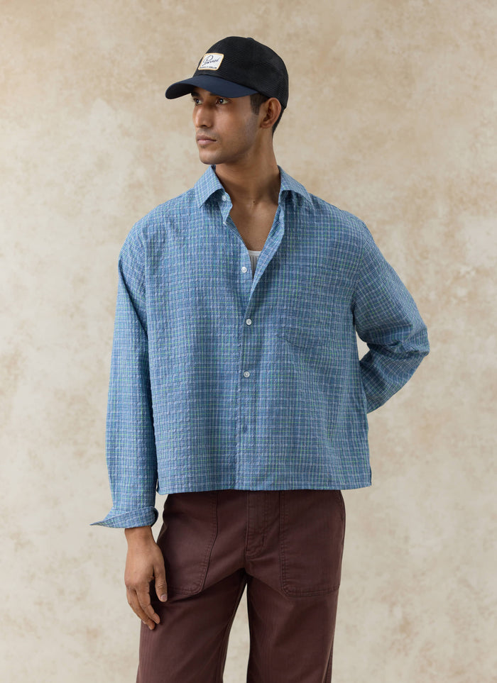 Model wears the blue Percival Hinsley Checked Boxy Shirt in cotton grid, styled with a navy trucker cap and brown trousers.