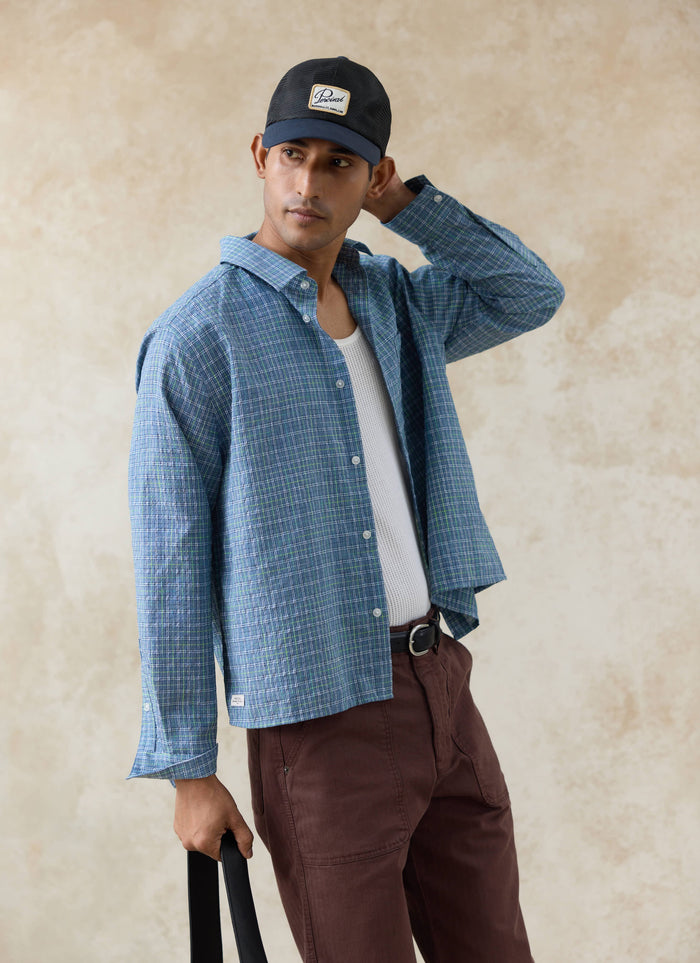 Model wears the blue cotton Percival Hinsley Checked Boxy Shirt and navy mesh trucker cap over a white mesh vest.
