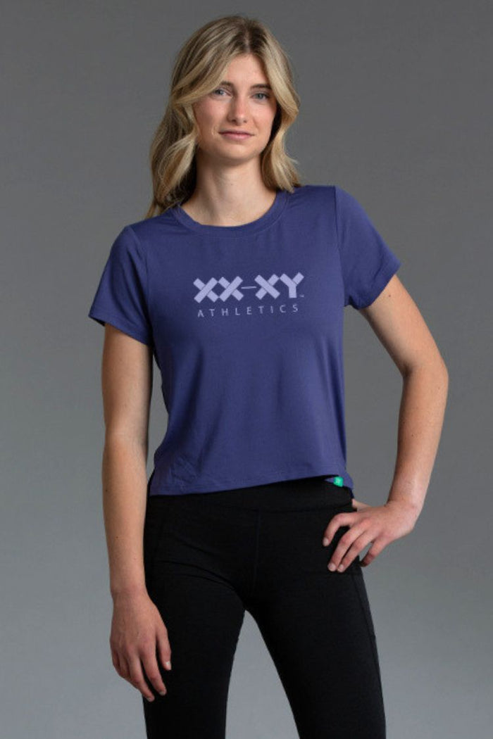 Woman wearing a blue 'XXXY Athletics' t-shirt on a gray background || Indigo