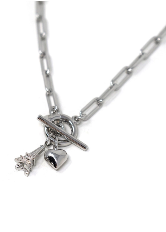 A silver paperclip chain necklace with a toggle clasp and two dangling charms: a small Eiffel Tower charm and a heart charm, shown on a white background.