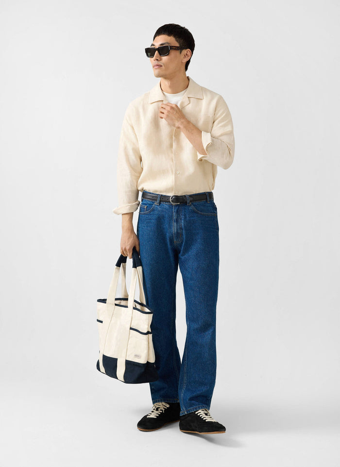 A model wears the Percival cream long sleeve cuban linen shirt, styled with dark blue jeans, sunglasses and a tote bag.
