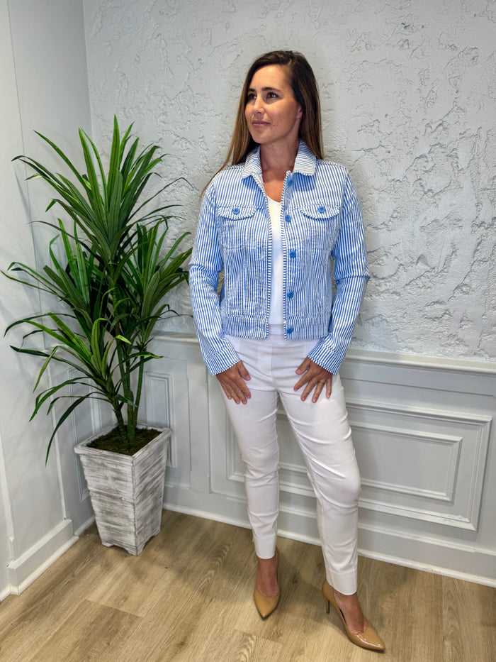 Smiling model in front of a white wall with a green plant in the corner. She's wearing the Lulu B Linen Travel Jacket - Seersucker, a light linen jacket with 2 front pockets and lightly frayed hems. Paired with a white shirt and white pants.