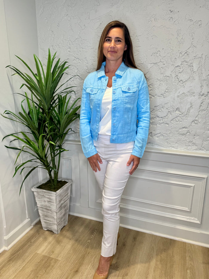 Smiling model in front of a white wall with a green plant in the corner. She's wearing the Lulu B Linen Travel Jacket - Clear Blue, a light linen jacket with 2 front pockets and lightly frayed hems. Paired with a white shirt and white pants.