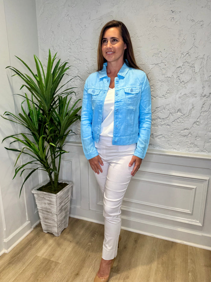 Smiling model in front of a white wall with a green plant in the corner. She's wearing the Lulu B Linen Travel Jacket - Clear Blue, a light linen jacket with 2 front pockets and lightly frayed hems. Paired with a white shirt and white pants.