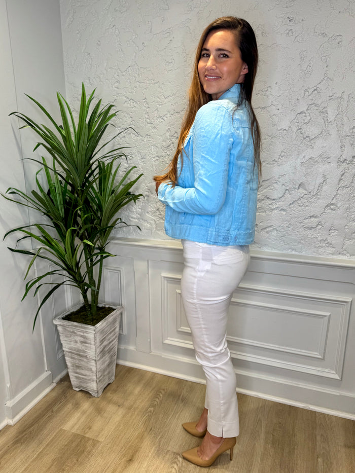 Side view of smiling model in front of a white wall with a green plant in the corner. She's wearing the Lulu B Linen Travel Jacket - Clear Blue, a light linen jacket with 2 front pockets and lightly frayed hems. Paired with a white shirt and white pants.