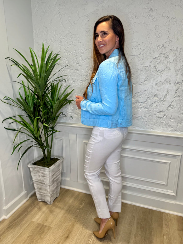 Smiling model in front of a white wall with a green plant in the corner. She's wearing the Lulu B Linen Travel Jacket - Clear Blue, a light linen jacket with 2 front pockets and lightly frayed hems. Paired with a white shirt and white pants.