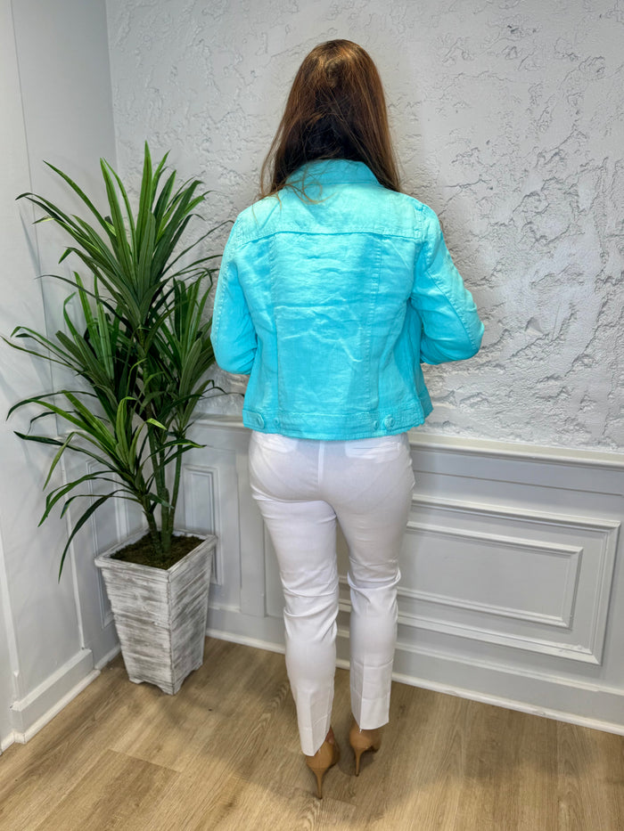 Back view of model in front of a white wall with a green plant in the corner. She's wearing the Lulu B Linen Travel Jacket - Seafoam, a light linen jacket with 2 front pockets and lightly frayed hems. Paired with a white shirt and white pants.