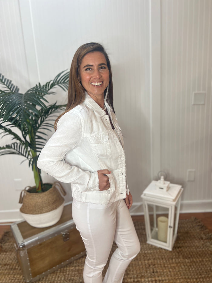A woman stands sideways with hand in pocket wearing a white linen jacket with a pearl necklace, white tank top and white pants with plant and antique trunk in the background