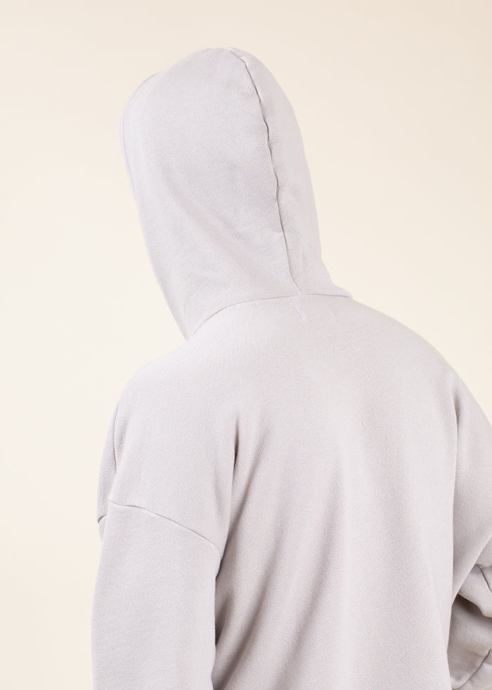 Person wearing Light gray hoodie with hood up facing away from the camera on a beige background