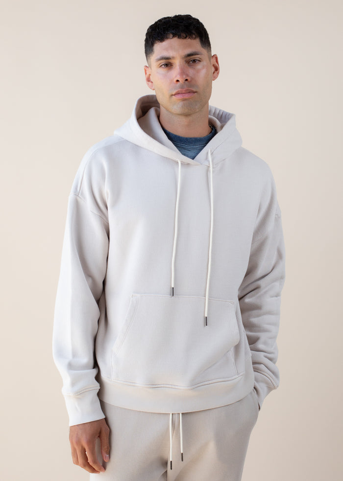 Man wearing an off white hoodie and beige pants against a plain background
