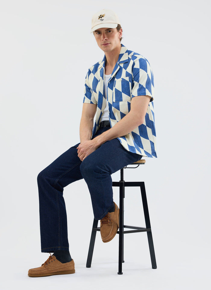 A model wears the Percival Market Diamond Cuban shirt in ecru and navy with dark jeans and a cap while sitting on a stool.