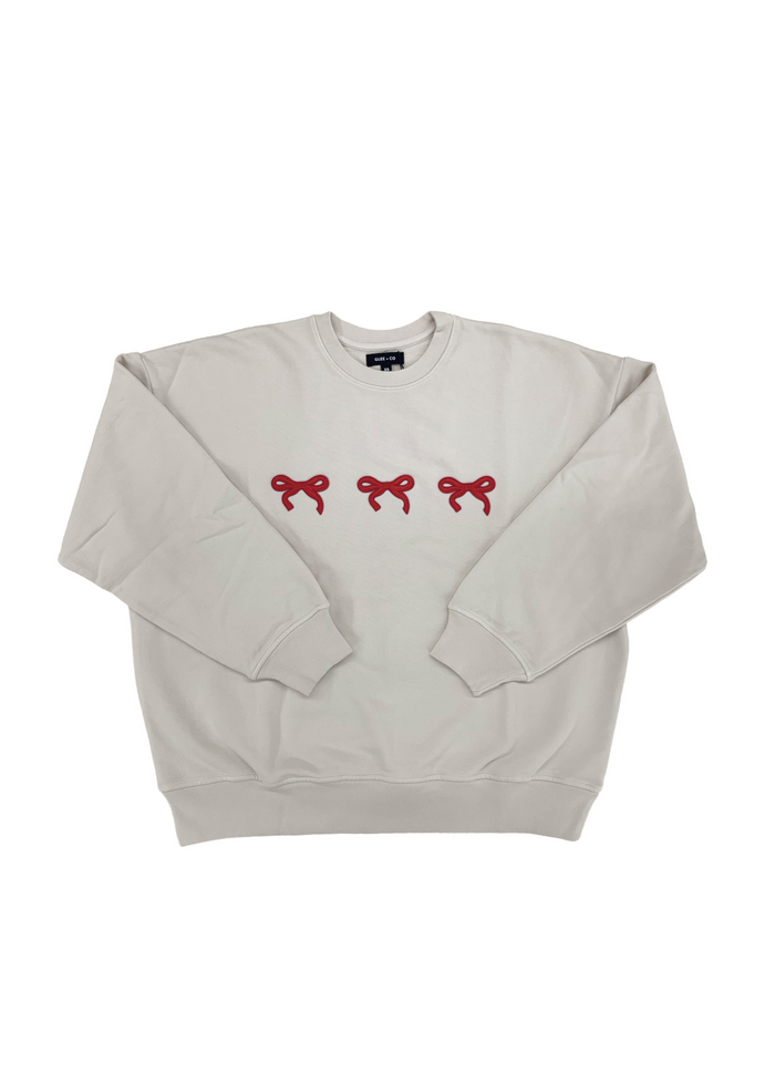 A light beige crewneck sweatshirt featuring three small red embroidered bows across the chest, displayed flat against a solid white background.