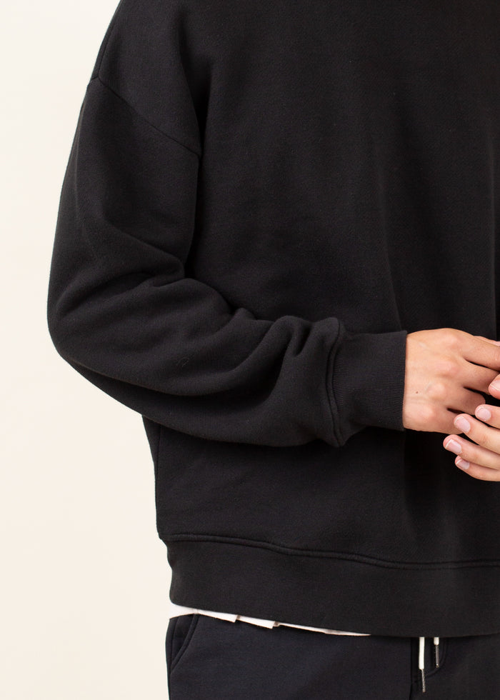Closeup of arm of Black crewneck sweatshirt worn by a person on a light beige background