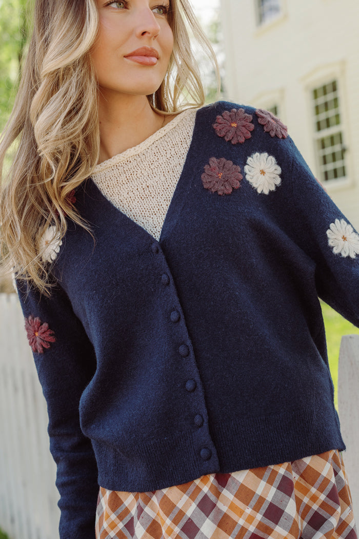 A woman wears the Ivy City Co Pauline Sweater in Midnight Floral—a button-front sweater featuring embroidered 3D floral embellishments in burgundy and cream—layered over a cream knit top and paired with a plaid skirt.