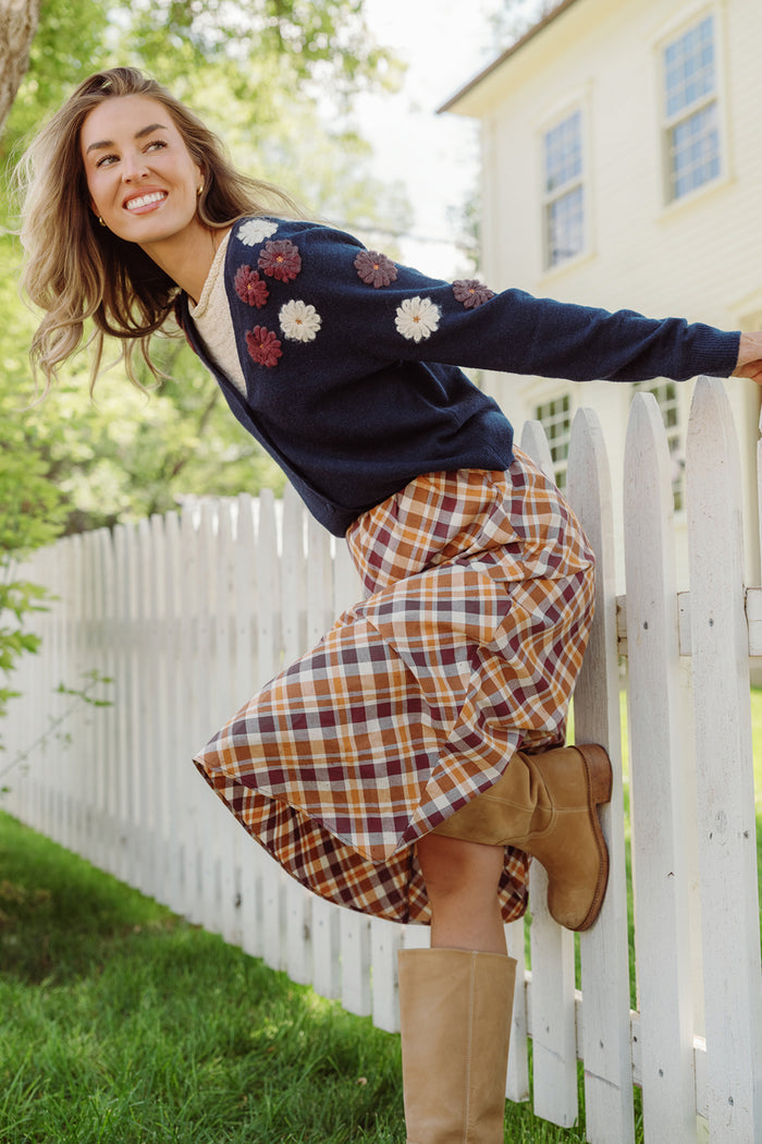 A woman is leaning on a white picket fence outdoors, smiling, and wearing the Ivy City Co Pauline Sweater in Midnight Floral paired with a plaid ruffle skirt and tall tan boots.