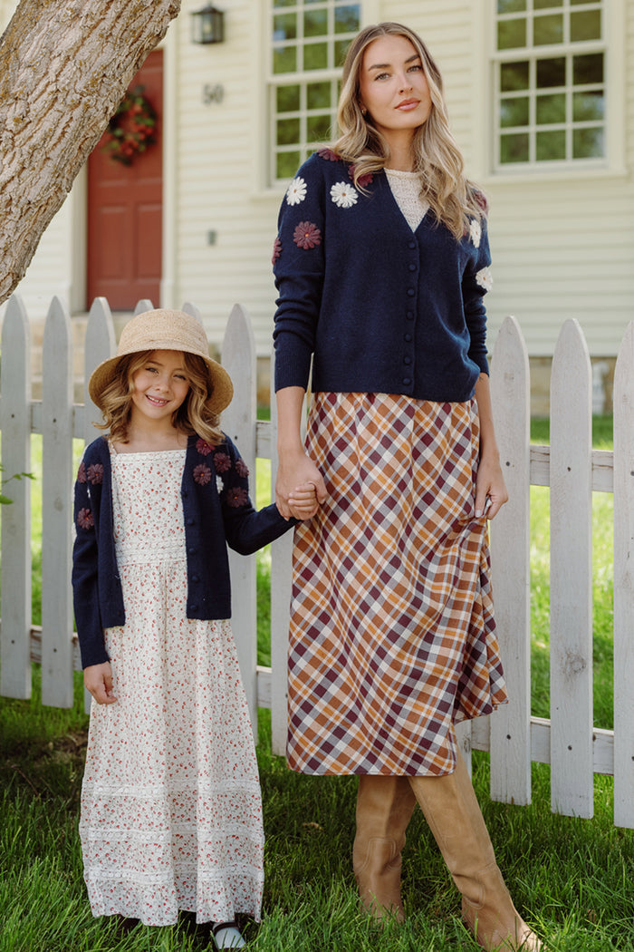 A woman and girl hold hands beside a white picket fence, both wearing the Ivy City Co Pauline Sweater in Midnight Floral. The woman completes her look with a plaid midi skirt, while the girl pairs hers with a floral maxi dress and straw hat.