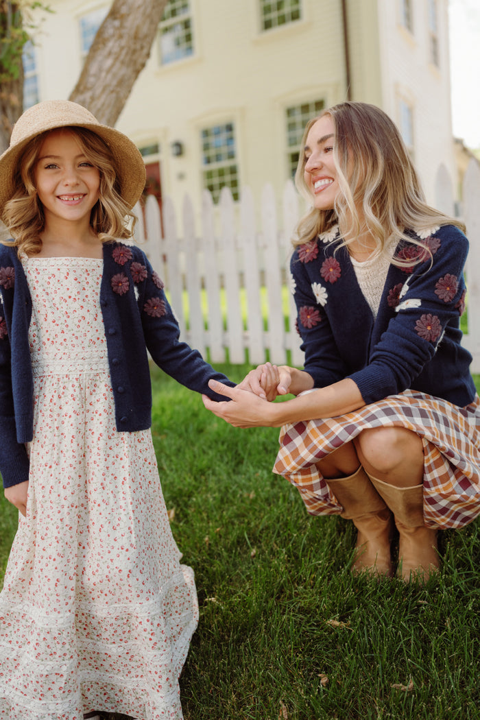 A woman kneels on the grass, smiling and holding hands with a young girl. Both wear matching Ivy City Co Pauline Sweaters in Midnight Floral. The girl pairs hers with a sunhat and white dress, while a white picket fence and house appear in the background.