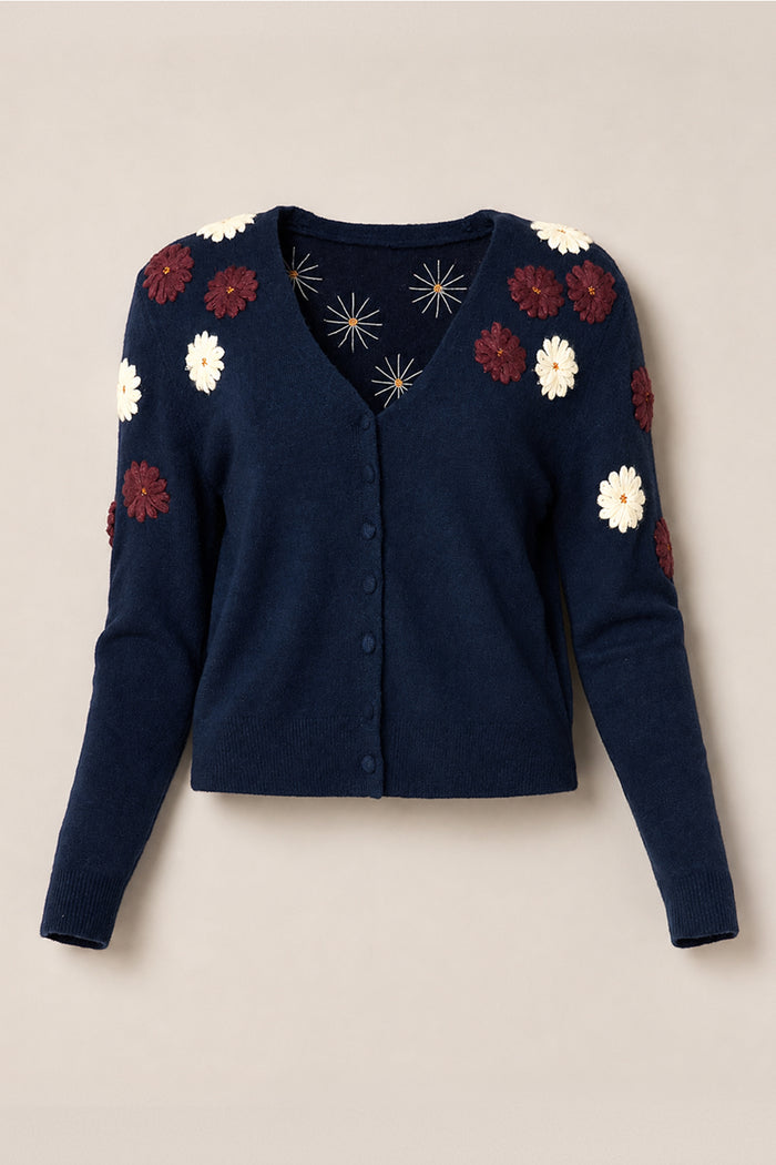 The Ivy City Co Pauline Sweater in Midnight Floral is a navy blue cardigan featuring long sleeves and white and burgundy floral appliqués on the shoulders and sleeves, shown against a plain beige background.