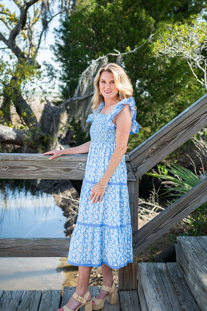 Woman in a blue dress standing on a wooden bridge by a body of water with trees in the background.