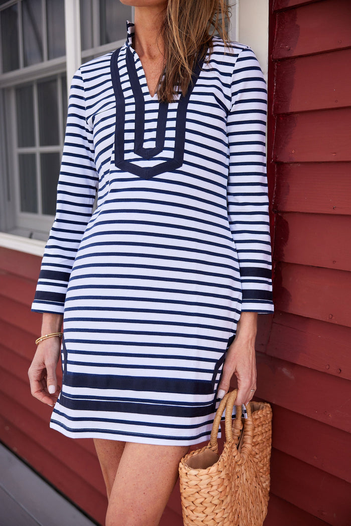 Model standing on a porch in front of a red building with big white windows. She's wearing a mini shift dress with navy and white stripes and navy piping around the collar, sleeve, and bottom hem