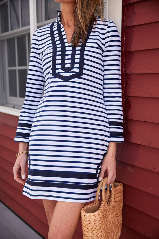 Model standing on a porch in front of a red building with big white windows. She's wearing a mini shift dress with navy and white stripes and navy piping around the collar, sleeve, and bottom hem