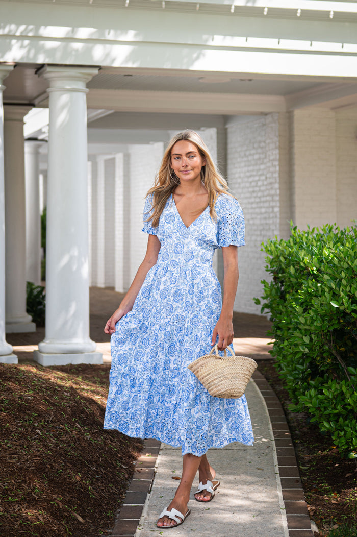 Woman in a blue floral dress holding a straw bag on a sunlit path.