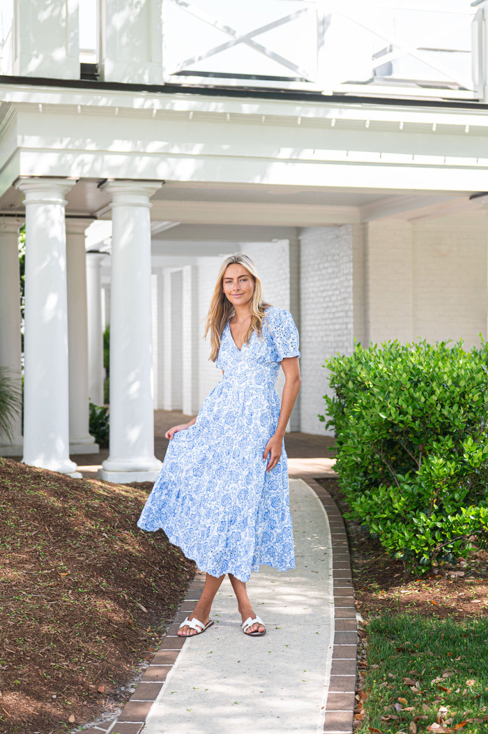 Woman in a blue dress standing on a pathway with white columns and greenery in the background
