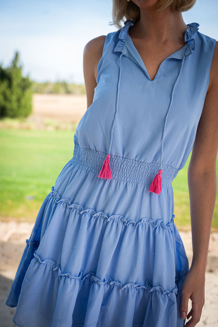 Model in a mini length, sleeveless periwinkle dress. It is ruched at the waist and tiered. It has pink tassels hanging from the collar.